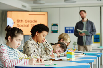 Side view at diverse group of children sitting in row and taking test in school classroom with...