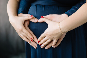Future parents keep their hands on the stomach of a young pregnant woman in the shape of a heart. The concept of love, care, future motherhood. Closeup view.