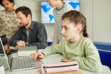 Side view portrait of cute schoolgirl using laptop in modern school classroom with kids in background
