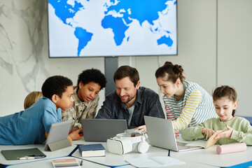 Front view portrait of smiling male teacher with diverse group of children using laptop together in...