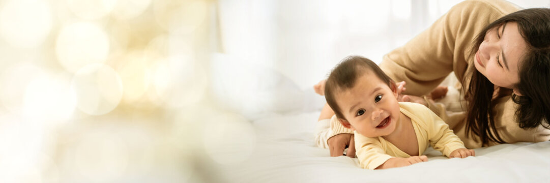 baby girl with asian mom smiling baby happily playing together in bed in the bedroom at home web banner with copy space on left