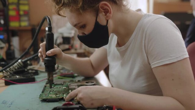 Female electronic repair service center worker repairs electronic boards and soldering microchip with heat gun. Women in mask. Technical occupations.