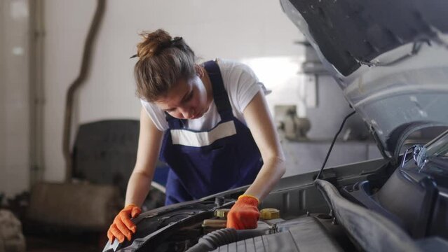 Female mechanic inspecting car liquids and pipes. Woman in overalls and protective gloves checks the engine and components at a car repair service. Vehicle failure fixing at modern workshop.
