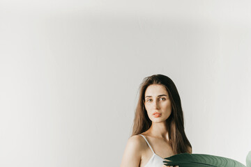 Portrait of an attractive young woman with dark long hair with a palm leaf in her hands against a gray wall. Copy space