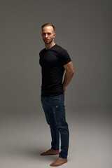 An attractive white man in jeans and black shirts tands in full growth against a gray background. Copy space