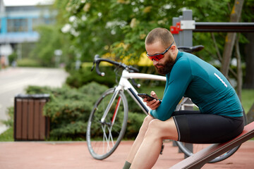 Obraz premium Stylish handsome guy cyclist rests on a wooden bench near bicycle in summer park.