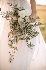 A bride in a white puffy dress close-up holds a wedding bouquet of wild flowers in white tones. Peonies, roses, petals.