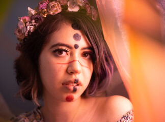 young woman with an alternative makeup or face paint or body art with a flowery dress and a flower crown staring at the camera with a warm lighting and a dreamy effect caused by an intentional blur