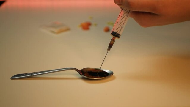 Close up of caucasian hand filling up syringe with black liquid heroin. Man drawing heavy drug from a spoon into syringe with other prohibited substances in background. Concept of heavy addiction
