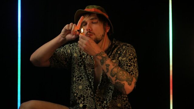 Slow motion shot of white dude in bucket hat smoking weed from a metal pipe. Close up of partying guy lighting up marijuana and inhaling it deeply. Concept of getting high from cannabis plants
