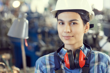 Portrait of smiling tomboy wearing hardhat and ear protectors standing at factory shop