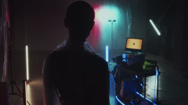 Rear silhouette of cyberpunk girl with shaved head in transparent waterproof coat standing in hideout with plastic film on walls, computers and neon lights