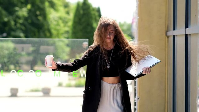 Happy young Caucasian businesswoman walking in city outdoors holding laptop and coffee-to-go while dancing listening to music in headphones. Female worker having fun finished working day