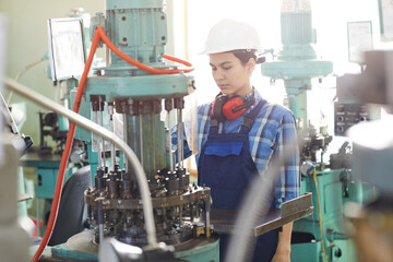 Serious young female technician in hardhat and overall standing in factory shop and working with...