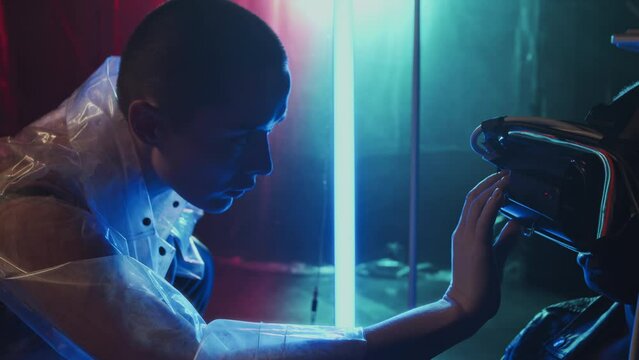 Side view of Caucasian cyberpunk girl with shaved head in transparent waterproof coat configuring VR goggles on Black man in hideout with plastic film on walls and neon lights