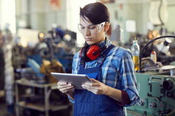 Serious young brutal woman in protective goggles and blue overall standing in workshop and reading technical specification on tablet