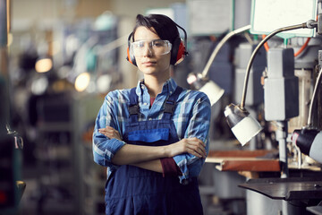 Portrait of serious professional female engineer in ear protectors and safety goggles standing at factory and crossing arms on chest