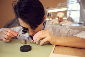 Close-up of focused young woman with short hair sitting at desk and looking through magnifying...