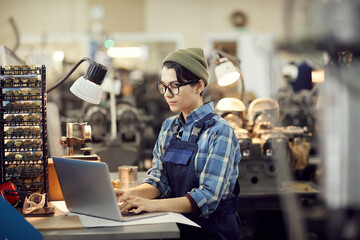 Serious skilled factory girl in eyeglasses sitting at table and making records on laptop while...