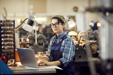 Portrait of content hipster female worker in eyeglasses sitting at desk and using laptop while...