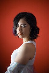 Portrait of a joyful fashionable young generation z woman wearing red lipstick and white eyeliner dressed in a white tank top and iridescent jacket happily posing in front of a red background