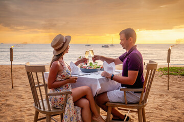 Couple in love drinking champagne on romantic dinner at sunset on the beach luxury restaurant. Young couple celebration Valentines Day, clink glasses at served table on sea sandy coastline.