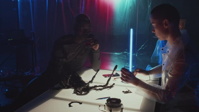Medium long slow motion of Caucasian woman with shaved head fixing robotic arm on body of Black man who sitting at table in front of her in dark room with neon lights, drinking and talking