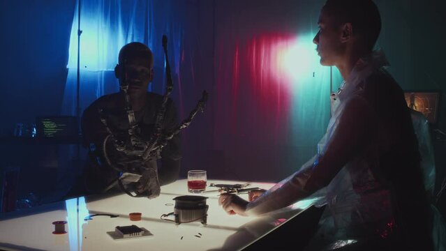 Medium long slowmo of cyberpunk girl with shaved head making robotic arm of young man who sitting at table in front of her in dark room with neon lights and covered with plastic film, admiring result