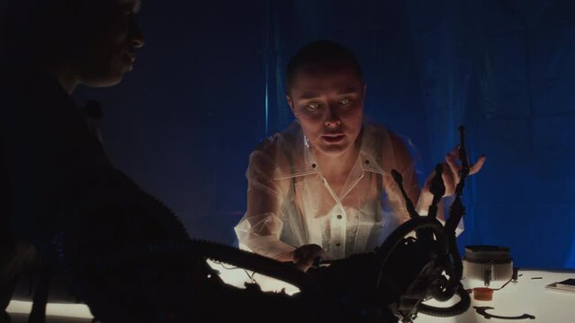 Medium slow motion of Caucasian cyberpunk girl with shaved head fixing robotic arm on body of Black man who sitting at table in front of her in dark room with neon lights, drinking and talking