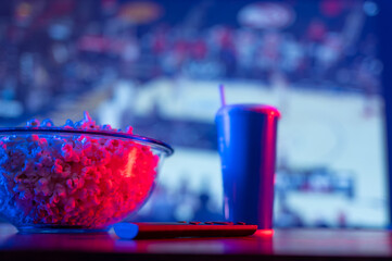 Stadium on the big TV screen. Popcorn in a bowl, a carbonated drink in a plastic glass and a remote...