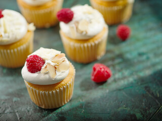 On a dark green background, sweet muffins with whipped cream, almonds and raspberries. Lots of objects. Restaurant, hotel, cafe, deli, confectionery. There are no people in the photo.