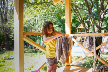 Cute child girl hangs clean linen on dryer outdoor, help with housework and children's duties at home, summer