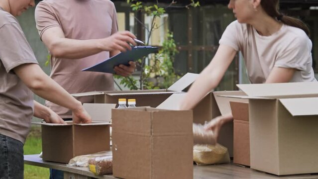 Group of young volunteers stacking food, groceries in donation boxes outdoors. Young people volunteering to sort donations for charity food drive. Volunteers packs canned goods and cereals in a