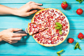 Woman's hands cut the homemade strawberry custard tart decorated with strawberry on blue wooden background. Top view