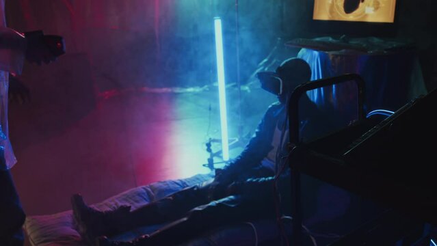Medium long of Cyberpunk girl in plastic checking on African American guy in VR glasses who connected to metaverse, sitting on floor in hideout with neon lights and smoke
