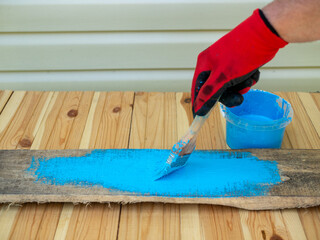 The process of painting wooden planks with blue paint. Hand holding brush painting blue stain on wood plank. The hand is wearing a red glove. Wooden background.