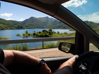 Conduciendo un coche con las manos al volante, mirando por la ventana un paisaje de naturaleza verde con montañas y un lago azul en el norte de España, verano de 2021