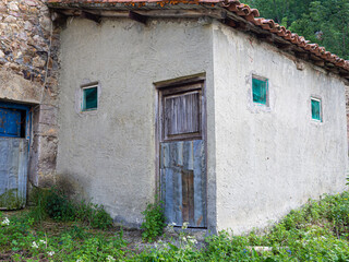 Fachada de una casa rural vieja, con una puerta desgastada y ventanas verdes en el campo, rodeada de hierbas verdes en Asturias, España, verano de 2021