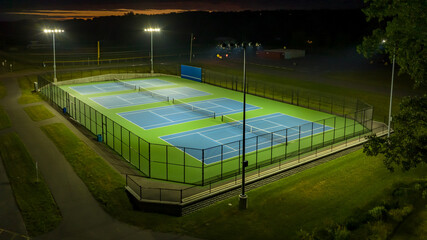Evening aerial photo of outdoor blue tennis courts with pickleball lines with lights turned on.	