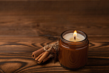 Obraz na płótnie Canvas Cozy burning candle in brown glass jar with sticks and cinnamon scent on brown texture wood.winter home decor.Soy ecological candle.Home and interior decoration.Place for text.Copy space.