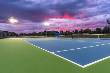 Evening photo of outdoor blue tennis courts with pickleball lines with lights turned on.	 - Powered by Adobe