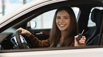 Happy young Caucasian woman showing key from auto, sitting inside car salon, buying new automobile at dealership, banner