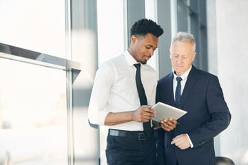 Fototapeta premium Two multi-ethnic business specialists in formalwear standing in lobby and using tablet while testing new app for data collection