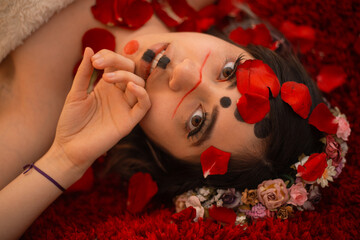 Close portrait of woman lying on a red furry textured rug with her face painted with an alternative makeup and with rose petals covering her face and a colorful flower crown in her hair - Powered by Adobe