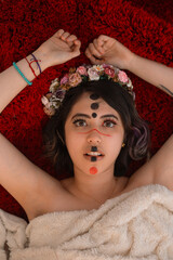 Close portrait of woman lying on a red furry textured rug with her face painted with an alternative makeup and with rose petals covering her face and a colorful flower crown in her hair