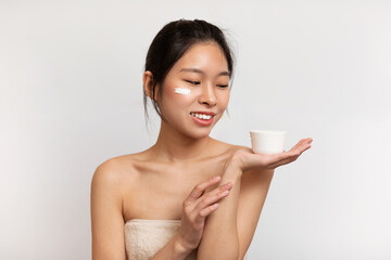 Glad asian lady with perfect skin applying moisturizing cream on face and holding jar with blank...