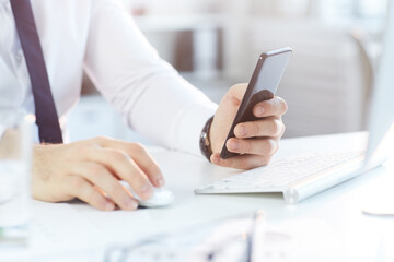Close-up of unrecognizable businessman in formal shirt sitting at desk and checking smartphone while working in office