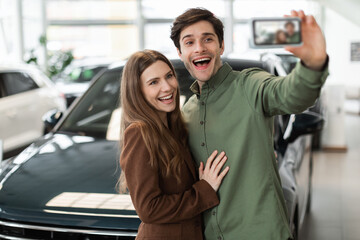 Excited young Caucasian couple taking selfie or filming video on smartphone near new auto at...