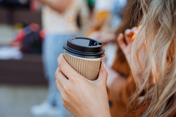 A glass of coffee is held by a girl's hand, a paper cup for hot drinks, wavy hair, food outside, a...