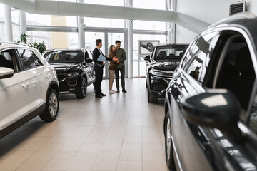 Full length of millennial couple choosing new car, having discussion with auto salesman at vehicle dealership